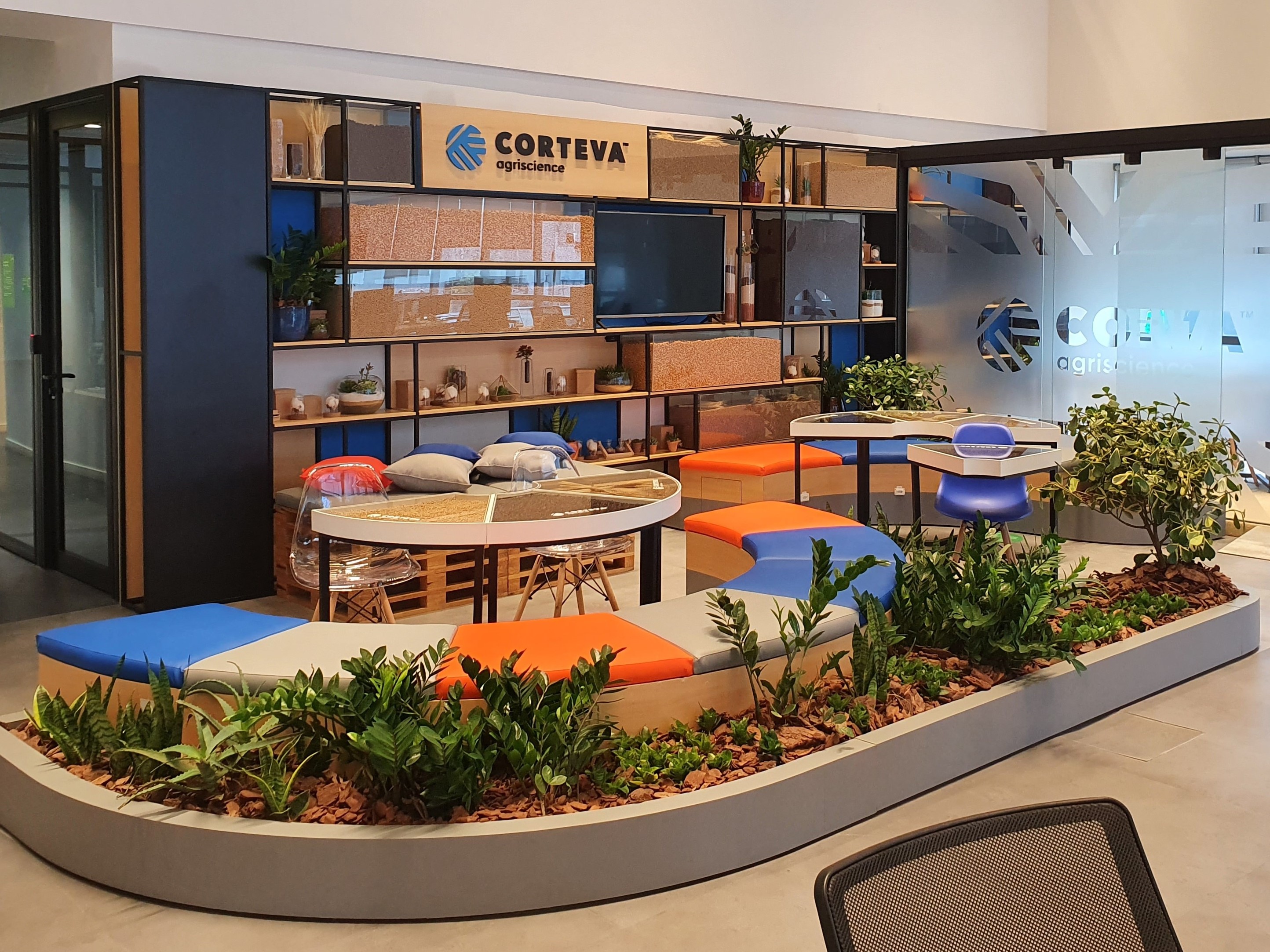 Workspace with tables and chairs in a circular shape in various colors and a panel in the background with various objects and the Corteva Agriscience logo.